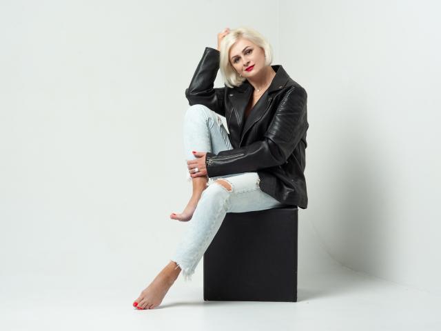 OliviaHorst with short blonde hair, red lipstick, black leather jacket, and ripped light blue jeans, sitting on a black cube, barefoot, hand in hair, confident, sexy.