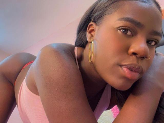 AvaBelle with dark skin, long black hair, gold earrings, nose ring, pink top, leaning forward, showing her ass, slightly parted lips, looking at the camera.