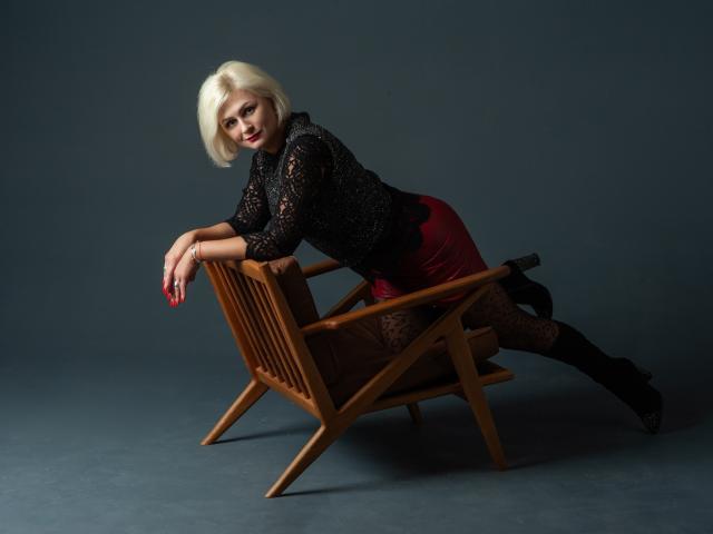 OliviaHorst with blonde bob, black lace top, red skirt, black lace tights, and boots, leaning on a wooden chair, showing her tits and ass, with a seductive look.