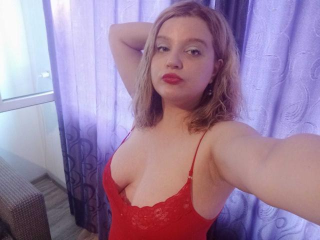 HospotsForU with blonde hair, red lipstick, and red lace lingerie, showing off her tits in a selfie, arms raised, in a room with purple curtains.