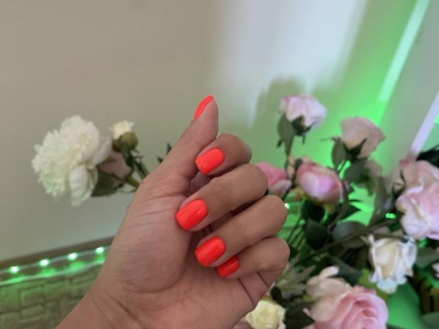 Trima's hand with bright neon pink nail polish, holding fingers together, showing off shiny, well-manicured nails. Pink roses and white chrysanthemums in the background.