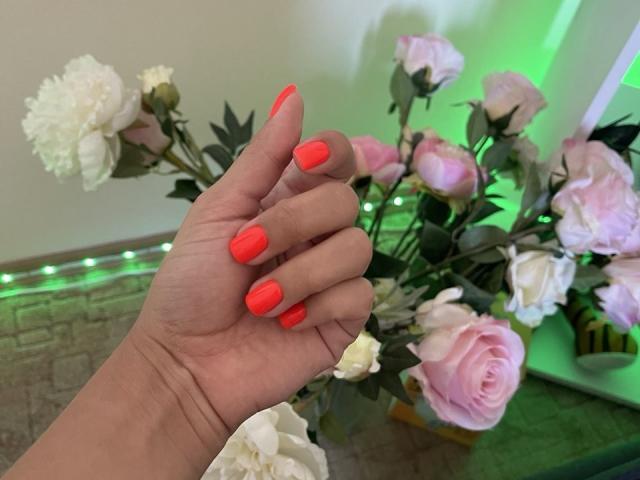 De hand van Trima, met fel oranje nagels, die haar manicure laat zien, naast een boeket roze en witte rozen. Haar huid is licht..