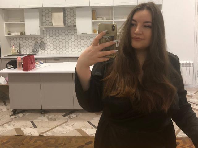 NataliePeterson with long brown hair, wearing a black dress, holding a phone in a white kitchen, taking a mirror selfie, showing her tits and a subtle smile.