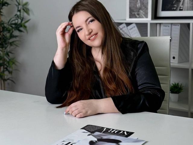 NataliePeterson, with long brown hair, black satin blouse, smiling, hand in hair, white desk, magazine with "A List" in front, white office chair, green plant in background.