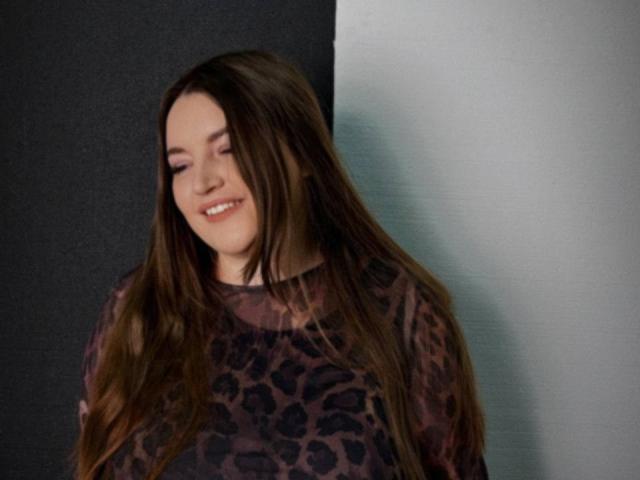 NataliePeterson with long brown hair, smiling, wearing a leopard print shirt, standing against a dark and light wall. Her eyes are closed, showing her cute nose.
