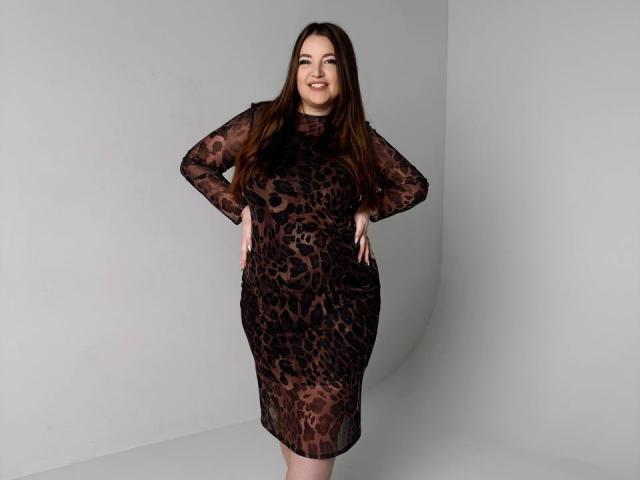NataliePeterson stands smiling, hands on hips, wearing a sheer black leopard print dress, showing her tits and ass through the fabric, with long brown hair.