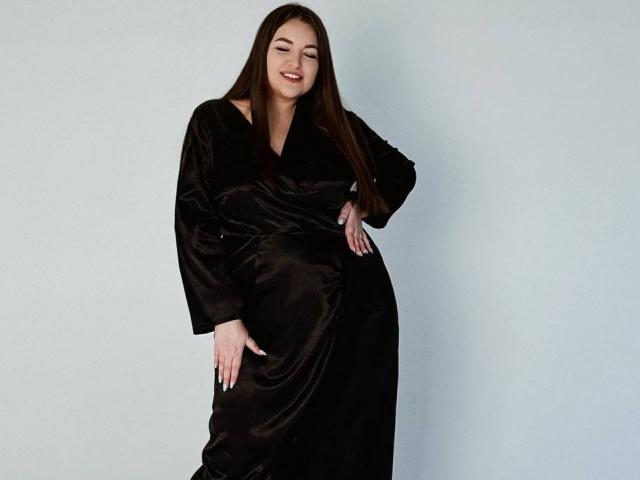 NataliePeterson, with long brown hair, smiling, wearing a black velvet dress, standing against a white wall, hand on hip, other hand touching her dress, showing her ass.