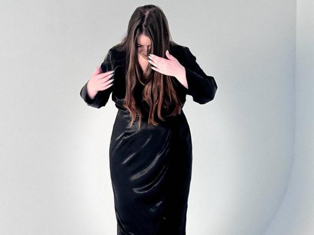 NataliePeterson with long brown hair, wearing a black dress, standing, hands near her face, looking down, soft light, white background, slim, black dress, gentle expression.