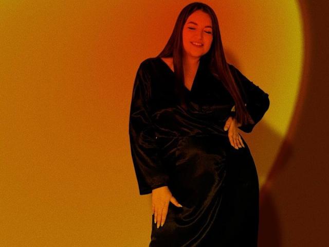 NataliePeterson with long brown hair, wearing a black silky robe, smiling, eyes closed, hand on hip, other hand on her ass, warm orange light, relaxed, confident.