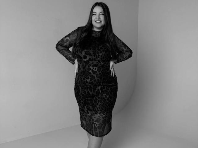 NataliePeterson with long dark hair, smiling, wearing a sheer black leopard print dress, hands on hips, standing, gray background, looking confident, curvy, sexy, full-ass, medium-boobs.