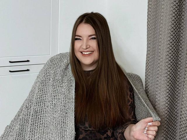 NataliePeterson with long brown hair, smiling, wrapped in a gray knitted blanket, wearing a dark patterned shirt, holding blanket on the right side.