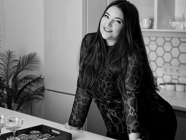 NataliePeterson with long dark hair, wearing a leopard print dress, leaning on a kitchen counter, smiling, showing her curvy ass, and a tray with glasses in front of her.