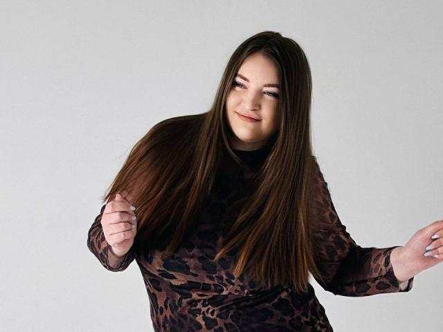 NataliePeterson with long brown hair, smiling, wearing a black leopard print top, hands raised, showing off her tits, against a plain white background.