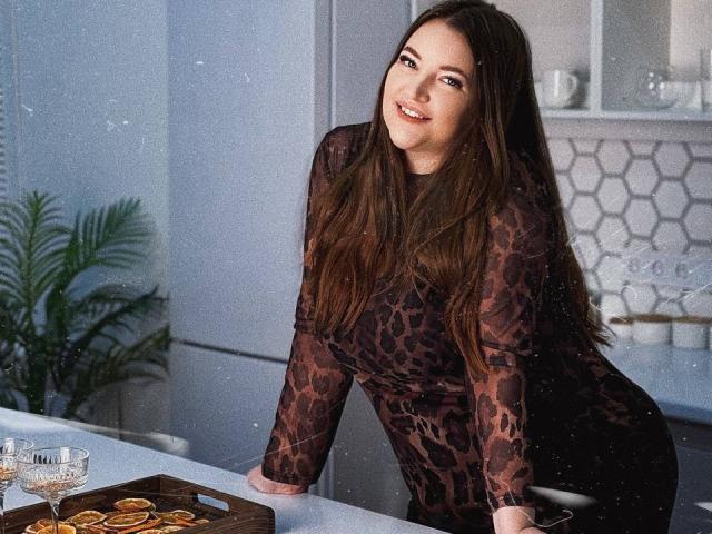 NataliePeterson with long brown hair, wearing a dark leopard-print dress, leaning on a kitchen counter, smiling, with a tray of dried orange slices and glass cups nearby.