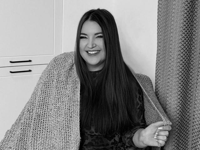 NataliePeterson with long dark hair, smiling, wearing a knitted blanket over a dark patterned dress, holding the blanket's edge, black-and-white photo, showing her cute, chubby face.