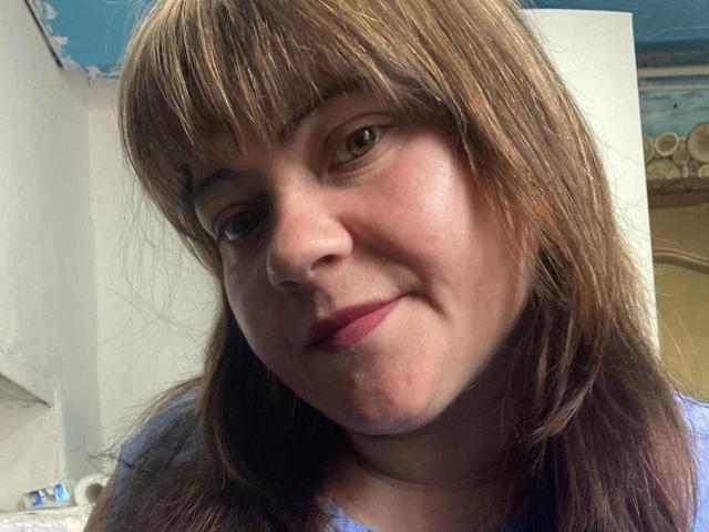 DebbieXRoy a les cheveux bruns clairs, lisses et avec une frange, une peau claire, des yeux marrons, et un léger sourire avec du rouge à lèvres rose. Elle porte une chemise bleue, et c'est un autoportrait..