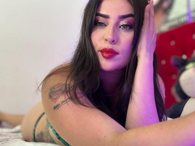 LindaSmith has long dark hair, red lipstick, and a tattoo on her arm. She's lying on a bed, looking at the camera with her hand on her head. Boobs visible.