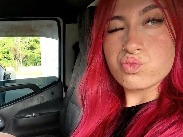 SammyViolet, com o cabelo rosa vibrante, piercing no nariz e lábios carnudos, piscando dentro de um carro, exibindo suas nádegas fartas e lábios cheios. Seus seios não estão visíveis..