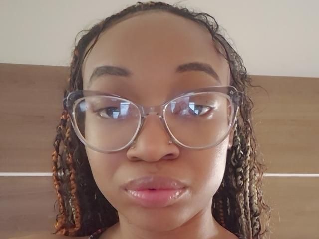 NadiaaSexy with brown skin, curly braids, clear glasses, nose ring, and pink lips, looking straight at the camera. Boobs not visible, focus on her face.