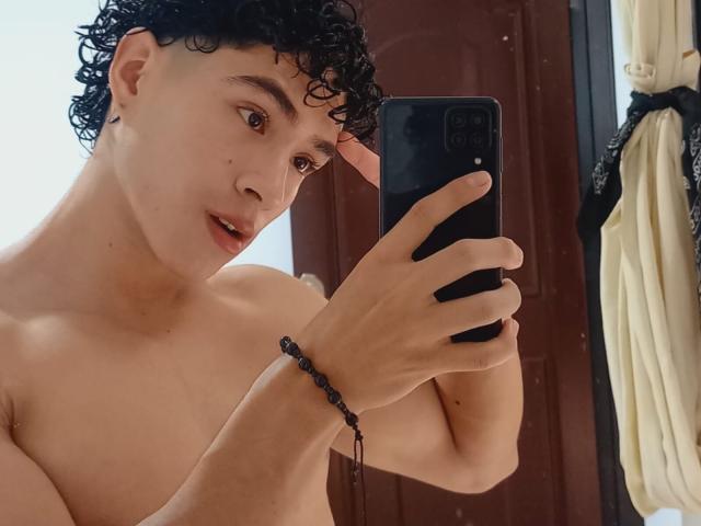 NoahJhoness with curly black hair, tan skin, and a bead bracelet, taking a mirror selfie, shirtless, holding a black phone, brown eyes, slightly open mouth.