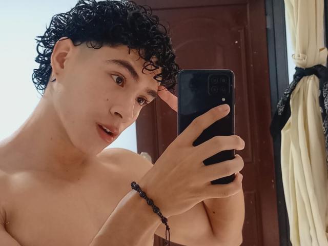 Shirtless NoahJhoness with curly black hair, brown eyes, and a bead bracelet, holding a phone, taking a mirror selfie. His lips are slightly parted.