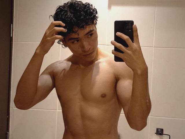 NoahJhoness, shirtless with curly black hair, holding a phone in a mirror selfie, showing a toned, tan-skinned body, small nipples, and a subtle smile.