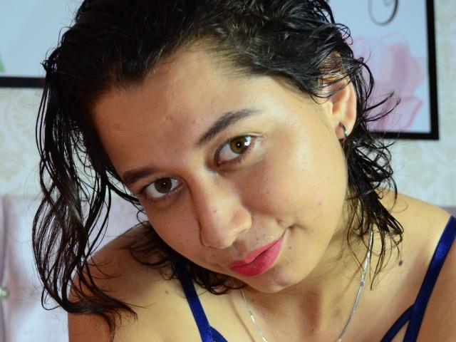 GalaSquirt has wet black hair, brown eyes, and is wearing a blue top. Her small tits and pink lips are visible. She's looking down with a slight smile.