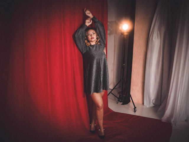 SallyVerda, met een sprankelende grijze jurk, armen omhoog, staand voor een rode gordijn, licht aan de rechterkant, hoge hakken, golvend blond haar, rode lippenstift, zelfverzekerde pose, studio omgeving..