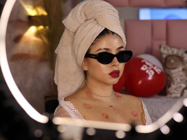 Kattana with a towel on her head, wearing black sunglasses, red lipstick, and a white lace top, has kiss marks on her chest. Boob tattoos, necklace, and ring.