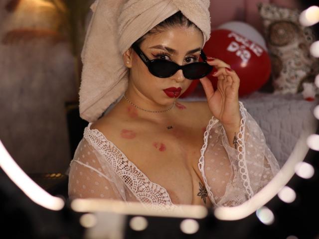 Kattana with a towel on her head, wearing black sunglasses, sheer lace top, red lipstick, and kiss marks on her chest, holding her sunglasses. Boobs visible.