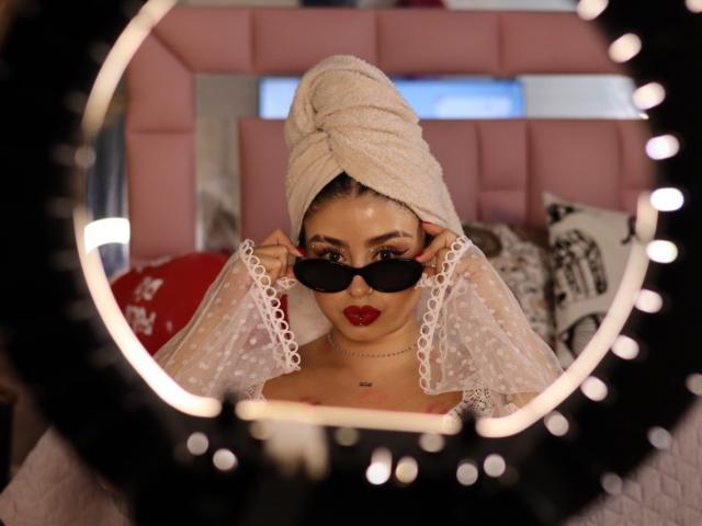 Kattana with a white towel turban, black sunglasses, and red lipstick, wearing a sheer white lace veil, reflected in a ring light. Her boobs are slightly visible.