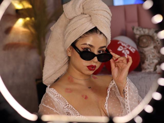 Kattana with a towel on her head, wearing black sunglasses, red lipstick, and a sheer lace top, has red lipstick marks on her chest and is touching her glasses.