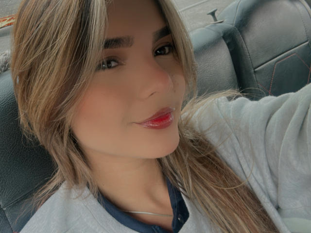 IvannaHazel's face, with red glossy lips, slightly parted, wearing a grey shirt, long brown hair, and a necklace, sitting in a car. Soft smile, light skin, brown eyes.