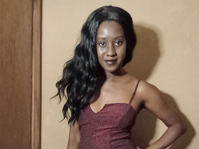 ChocolateSipp with dark skin, long wavy black hair, wearing a sparkly maroon dress, standing against a beige wall, smiling, hands on hips, looking at the camera.