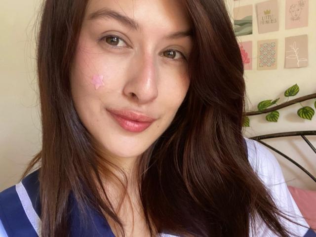 IsabellaPalacio has brown hair, pink nose sticker, and slight smile. She's wearing a blue and white top, with pink lips and brown eyes. Her ass is not visible.