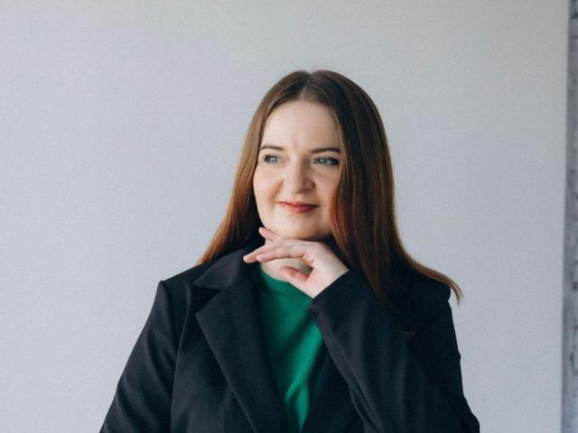 FreyaDak has long brown hair, blue eyes, and a slight smile. She's wearing a black coat over a green top, with her hand gently touching her chin.