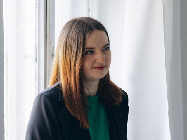 FreyaDak with light brown hair, green shirt, and black jacket, smiling slightly, standing by a window with white curtains, looking to the left.