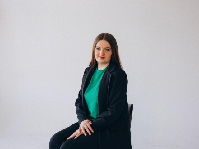 FreyaDak with long brown hair, green shirt, black jacket, and black pants, sitting on a chair, looking at camera, hands on lap, slight smile, soft natural light.