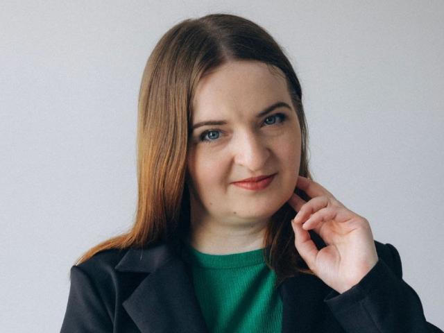 FreyaDak has light brown hair, blue eyes, and a slight smile, wearing a green sweater with a black coat, touching her chin with her right hand.