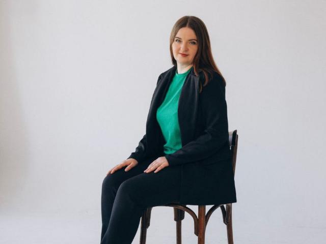 FreyaDak with long brown hair, green top, black blazer, and black pants, sitting on a wooden chair, smiling slightly, hands on her knees, white background.