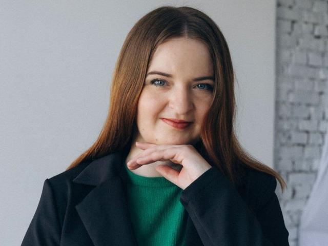 FreyaDak with long brown hair, blue eyes, and a slight smile, wearing a black coat over a green sweater, resting her chin on her hand.