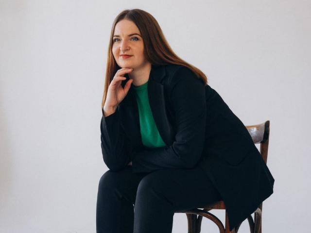 FreyaDak, con cabello largo y castaño, una blusa verde, un blazer negro y pantalones negros, sentada en una silla de madera, con la mano en la barbilla, sonriendo sutilmente, luciendo relajada y segura de sí misma..
