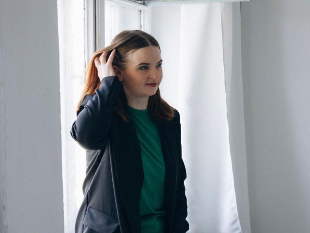 FreyaDak with light brown hair, wearing a green shirt and black jacket, standing by a window, hand in hair, smiling slightly, soft natural light, casual setting.
