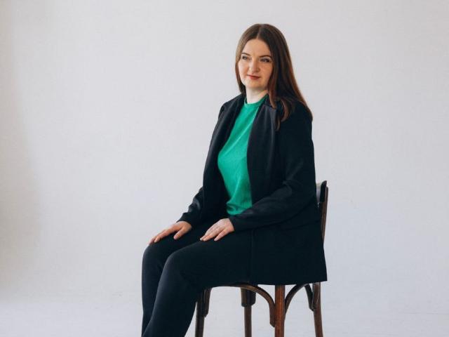 FreyaDak está sentada en una silla de madera, vestida con un blazer negro, una blusa verde, pantalones negros y calcetines negros, con el pelo largo y castaño y una ligera sonrisa..