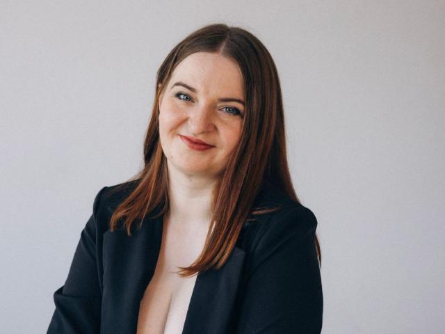 FreyaDak hat braunes Haar, blaue Augen und ein leichtes Lächeln. Sie trägt einen schwarzen Blazer mit tiefem Ausschnitt, der ihre Brüste zeigt. Sie hat helle Haut und ein hübsches Gesicht..