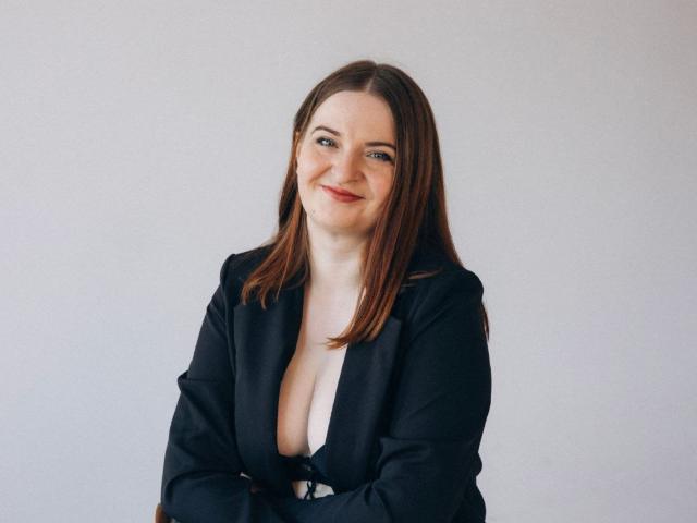 FreyaDak, with brown hair, smiling, wearing a black blazer, exposing her tits, arms crossed, sitting, white background.