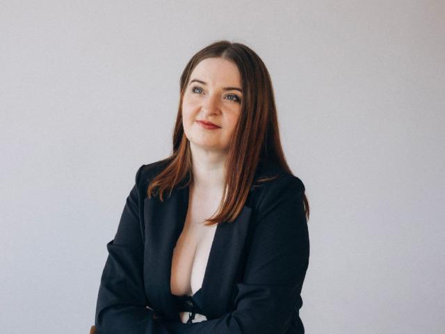 FreyaDak with brown hair, blue eyes, and a slight smile, wearing a black blazer with a deep cleavage, arms crossed, standing against a plain white background.