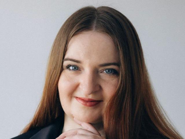 FreyaDak a les yeux bleus, les cheveux bruns et lisses, et un sourire discret, avec sa main sous son menton. Elle porte une chemise noire, et elle a l'air mignonne et détendue..