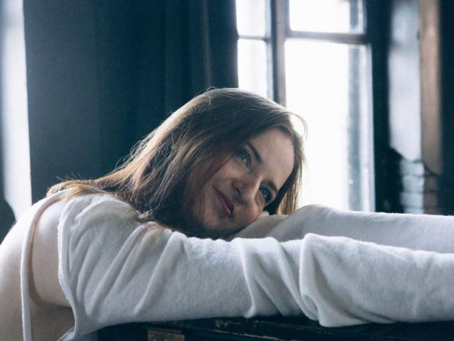 FreyaDak with long brown hair, blue eyes, and a slight smile, leaning on a dark surface, wearing a white long-sleeve shirt, with sunlight in the background.