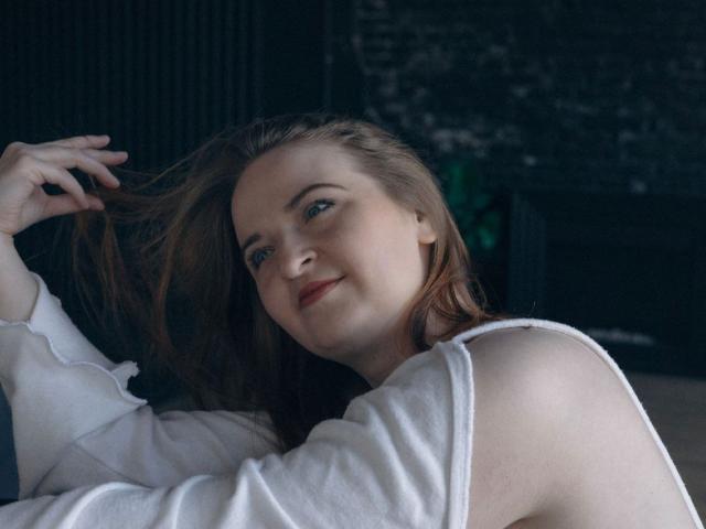 FreyaDak with light brown hair, blue eyes, and red lipstick, smiling, wearing a white shirt, holding her hair with one hand, soft light, relaxed expression.